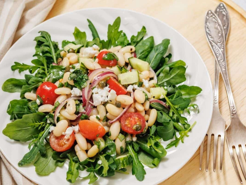 A fresh White Bean Salad on a white plate featuring arugula, white beans, cherry tomatoes, sliced red onions, avocado, and cheese, placed on a wooden surface with two forks beside it.