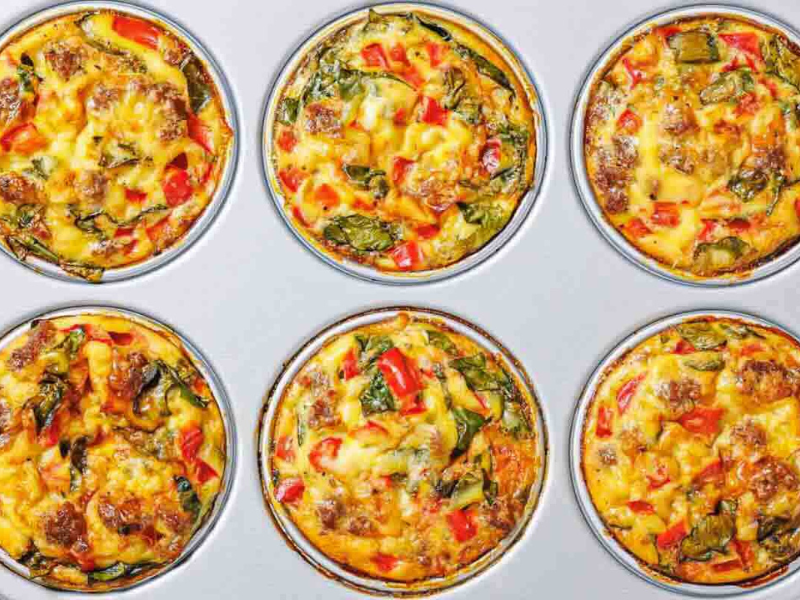 Six baked egg muffins with visible pieces of sausage, spinach, and red bell peppers in a muffin tin.