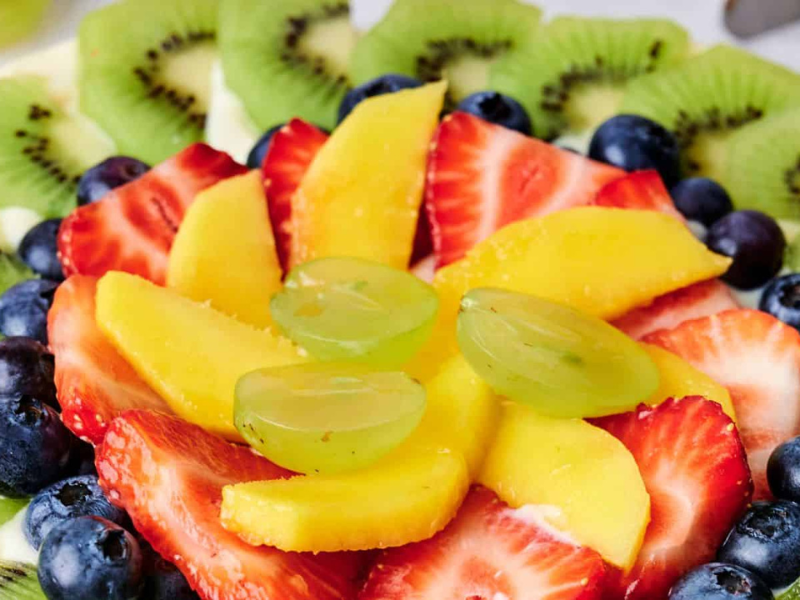 A close-up of a vibrant fruit tart topped with slices of kiwi, strawberries, mango, grapes, and blueberries arranged in a decorative pattern.