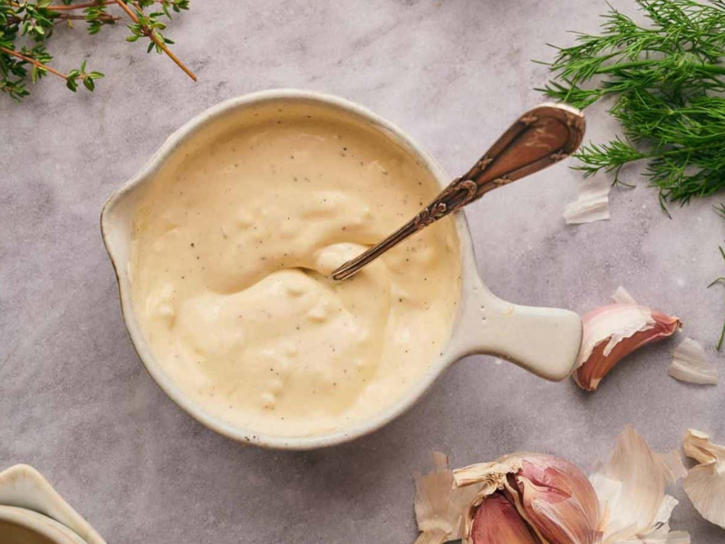 Garlic aioli in a mixing bowl with a spoon in it, garlic cloves, and dill around it.