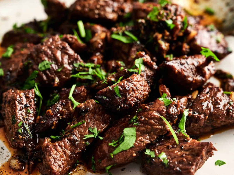 Close-up of cooked beef chunks garnished with chopped herbs on a plate with a spoon nearby.