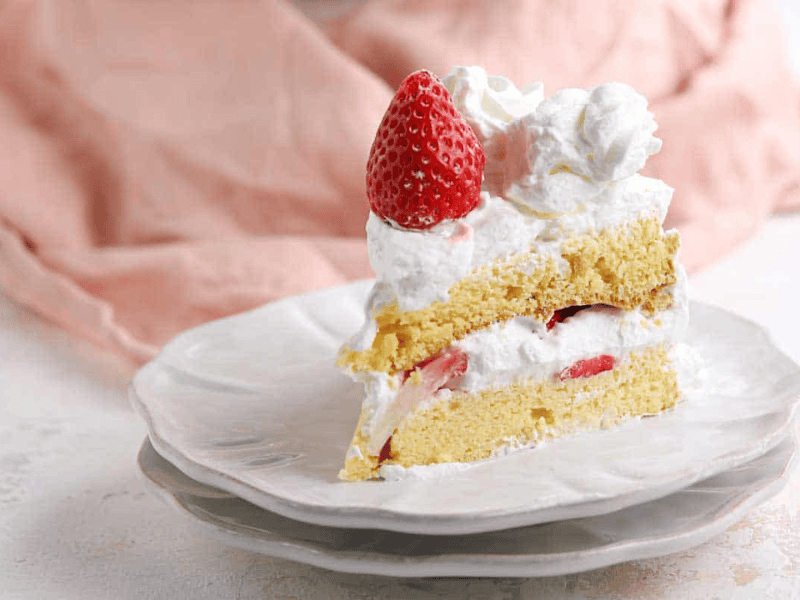 A slice of strawberry shortcake with whipped cream on a white plate, topped with a whole strawberry.