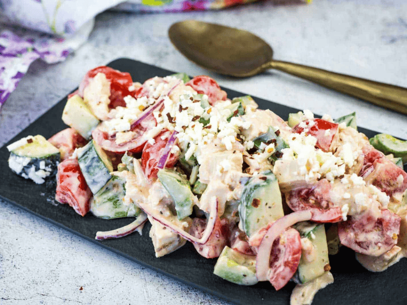 A plate of Greek Salad with Chicken sits on a dark serving tray next to a spoon.