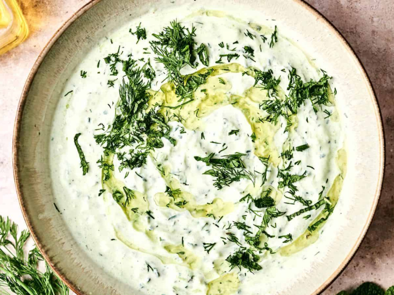 A bowl of creamy tzatziki sauce garnished with chopped dill and a swirl of olive oil. Fresh dill and garlic are nearby.