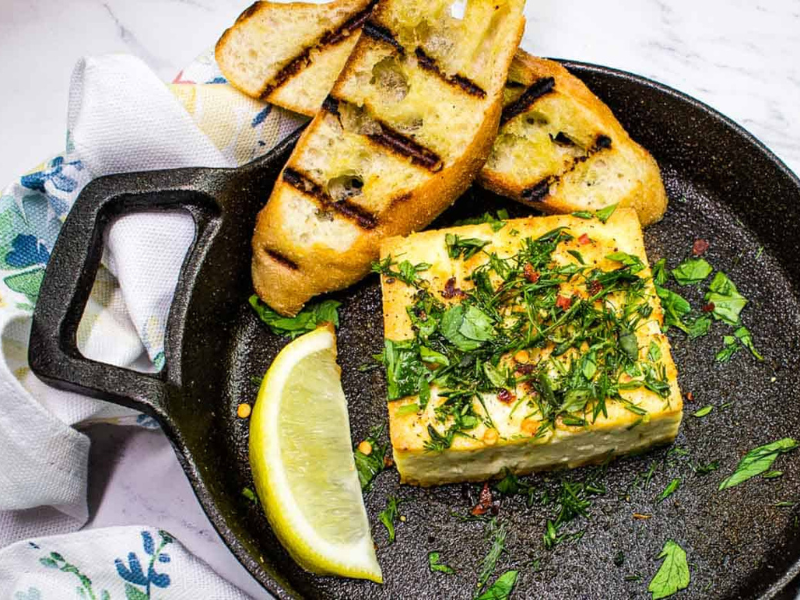 Grilled Feta with Herbs in a black serving dish with toasted bread and a lemon wedge.