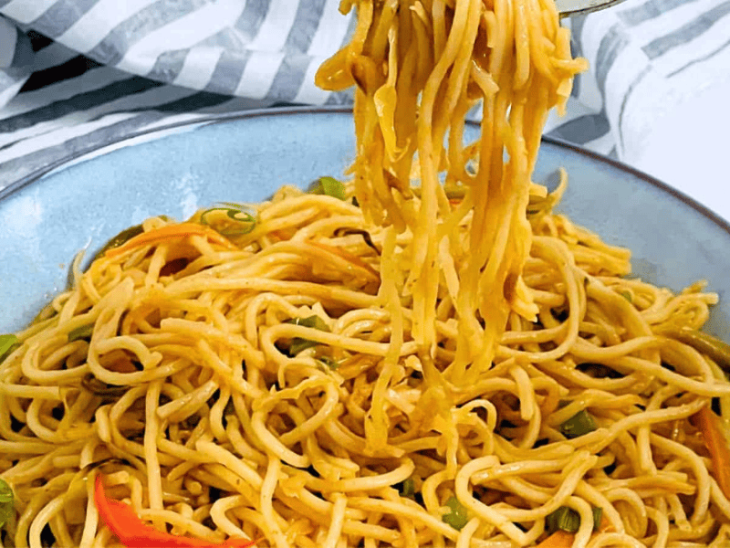 A bowl of Hakka Noodles with vegetables, a fork lifting some noodles, placed on a blue dish with a striped napkin on the side.