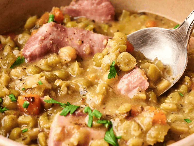 A bowl of split pea soup with chunks of ham, diced carrots, and herbs, with a spoon resting inside.