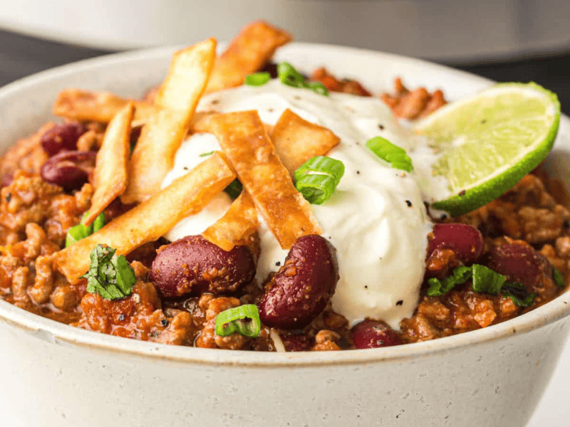 A bowl of chili topped with sour cream, crispy tortilla strips, sliced green onions, and a lime wedge.