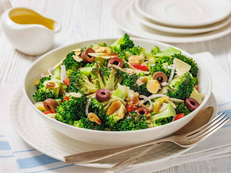 A bowl of Italian broccoli salad with sliced olives, almonds, onions, and red peppers, served on a white plate with a fork and napkin beside it.