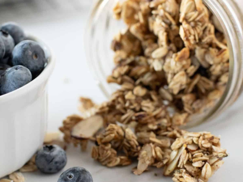 A glass jar of granola spills onto a surface next to a white bowl filled with blueberries.