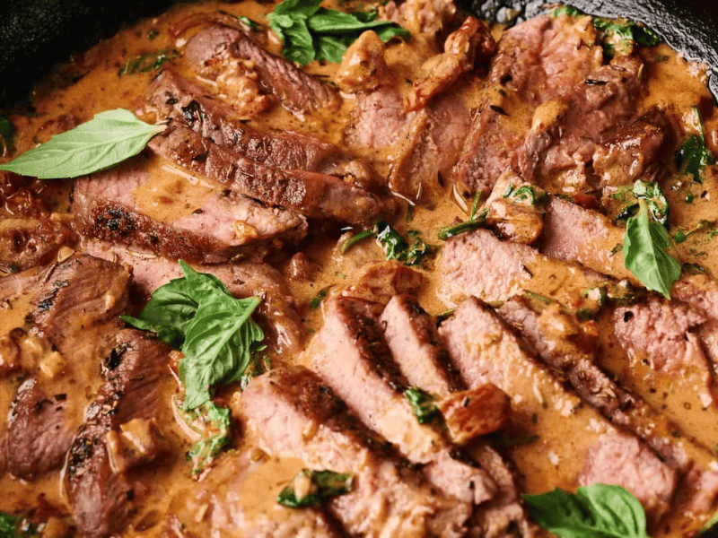 Slices of steak arranged in a creamy sauce with herbs and garnished with fresh basil leaves in a black skillet.