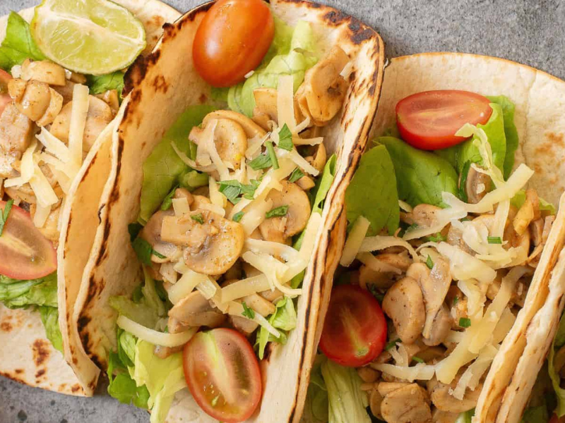 Three soft tacos filled with lettuce, mushrooms, shredded cheese, and cherry tomatoes, garnished with a lime wedge.