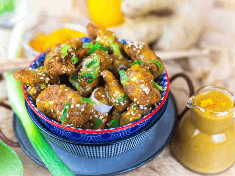 A bowl of fried chicken pieces coated in sauce and garnished with sesame seeds and green onions, accompanied by a small jug of sauce.