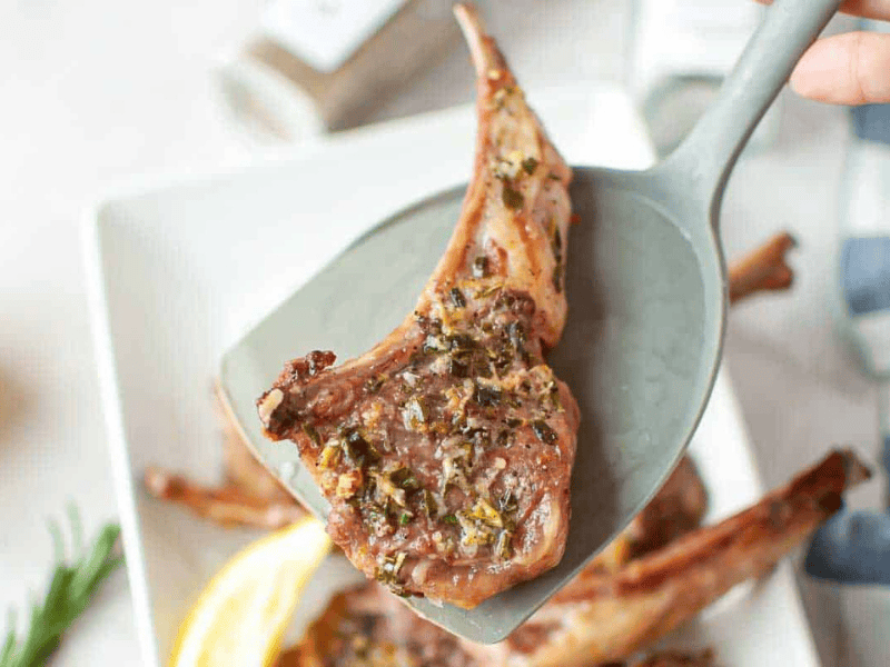 A cooked lamb chop seasoned with herbs is lifted on a gray spatula above a white plate with more lamb chops and lemon slices.