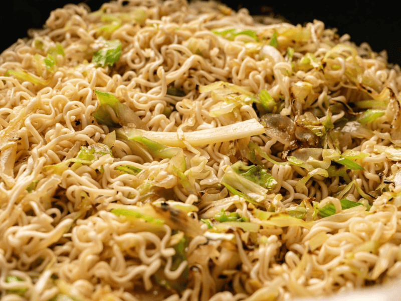 A close-up of cooked instant noodles mixed with saut&eacute;ed cabbage and onions in a pan.