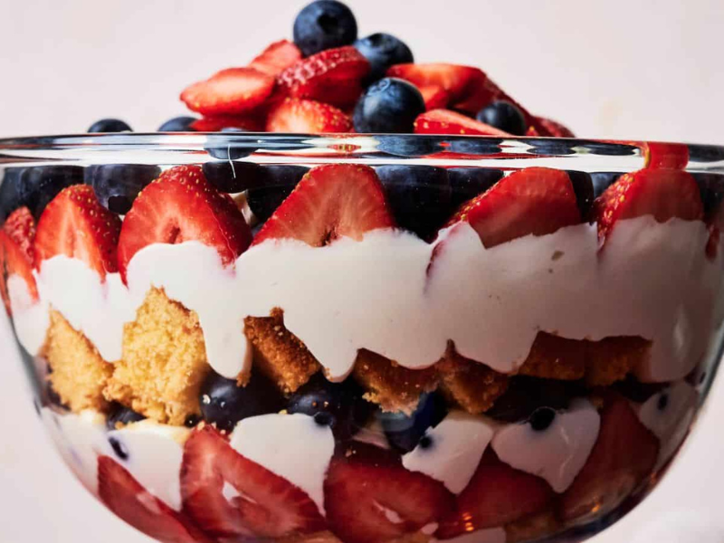 A glass bowl filled with layered dessert, featuring sliced strawberries, blueberries, whipped cream, and cake pieces.