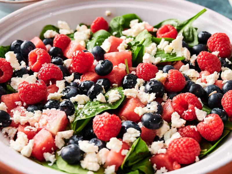 A salad with spinach leaves, watermelon cubes, blueberries, raspberries, and crumbled feta cheese in a white bowl.