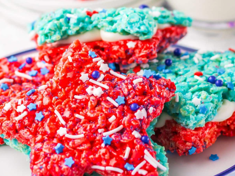 Patriotic Rice Crispy Treats in star shapes, decked out in red, white, and blue colors, are sprinkled with fun decorations and beautifully stacked on a plate.