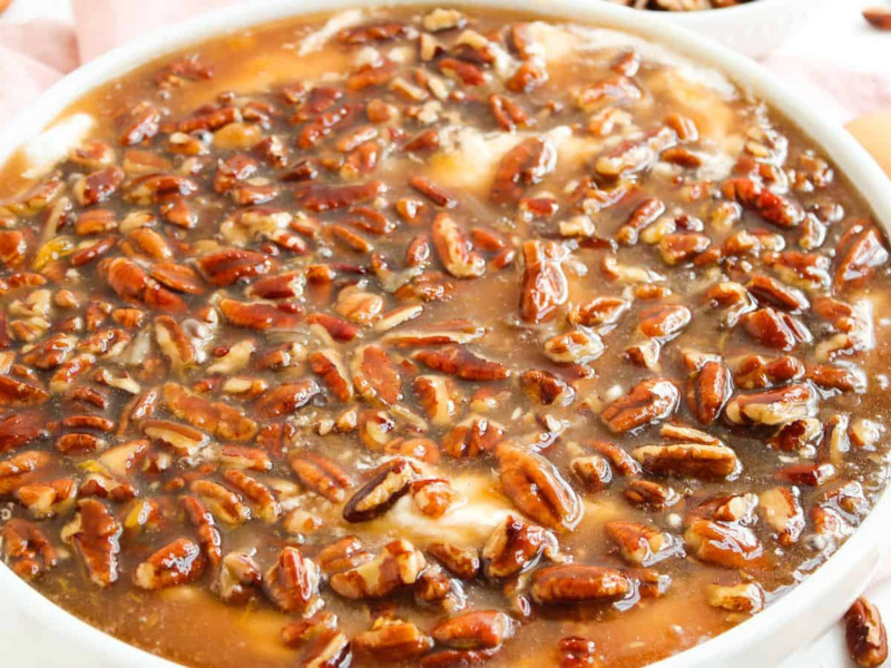A close-up of a dessert topped with a glossy layer of pecans and caramel sauce in a white dish, with vanilla wafers on the side.