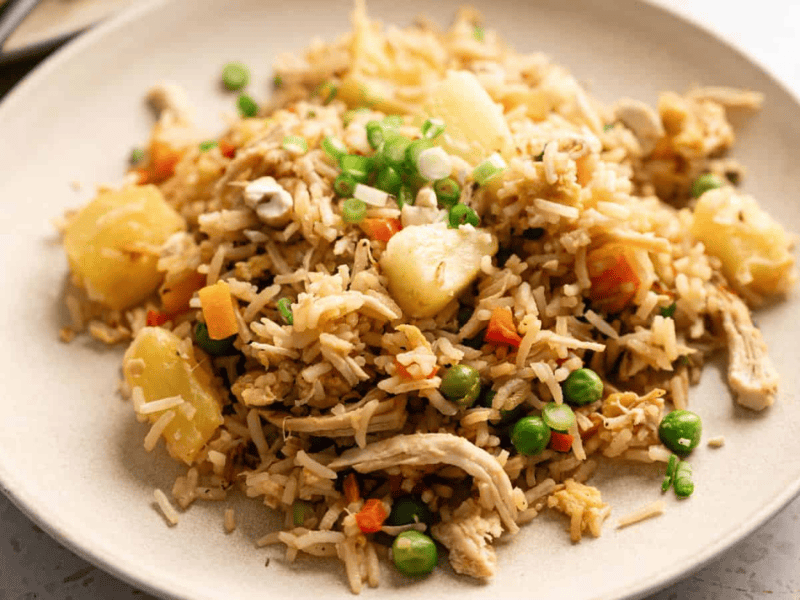 A plate of fried rice with peas, carrots, pineapple chunks, shredded chicken, and garnished with chopped green onions.