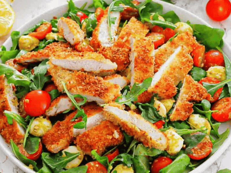 A plate of salad with arugula, cherry tomatoes, mozzarella balls, and sliced breaded chicken cutlets on top.