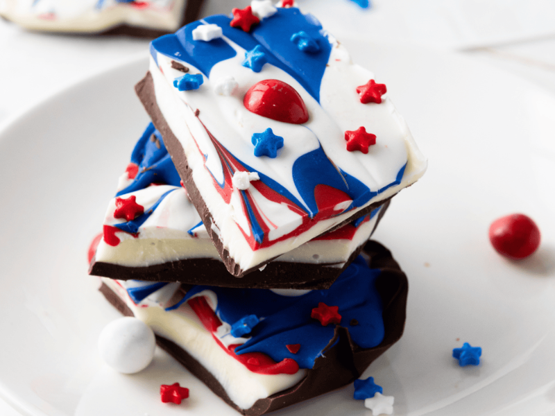 Three pieces of chocolate bark with red, white, and blue swirled topping and star-shaped sprinkles stacked on a white plate.