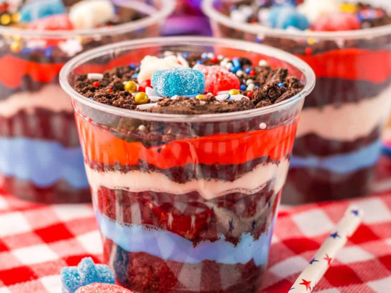 Three-layered red, white, and blue dirtcups with vibrant fillings are topped with various candies and sprinkles, all perfectly arranged on a red and white checkered cloth.