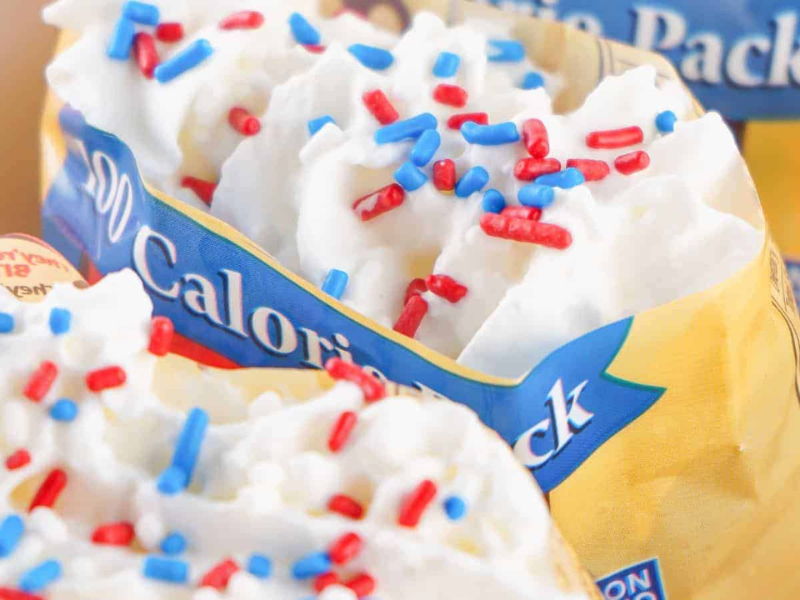 Close-up image of a snack pack filled with whipped cream topped with red, white, and blue sugar sprinkles. Packaging displays 
