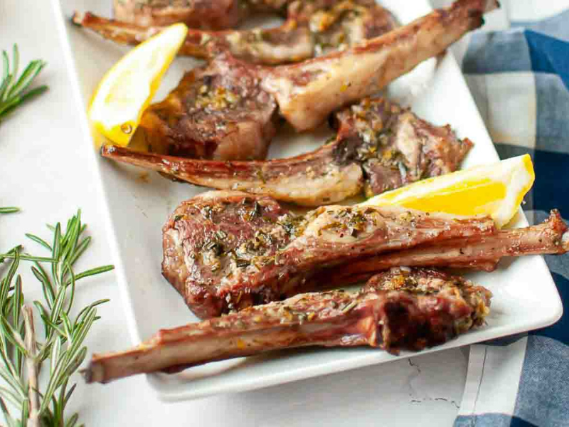 Grilled lamb chops garnished with herbs, served on a white rectangular plate with lemon wedges.