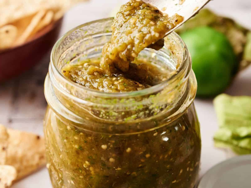 A jar of green salsa with a spoon lifting some out; tortilla chips and a tomatillo are visible in the background.