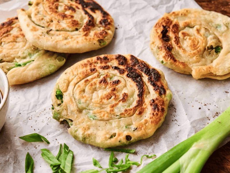 Four cooked scallion pancakes on parchment paper, garnished with sliced green onions, with a small bowl of dipping sauce and a whole scallion beside them.