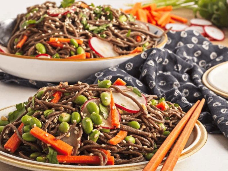 Two bowls of soba noodle salad with carrots, edamame, and radish slices, placed on a patterned cloth with wooden chopsticks beside them.
