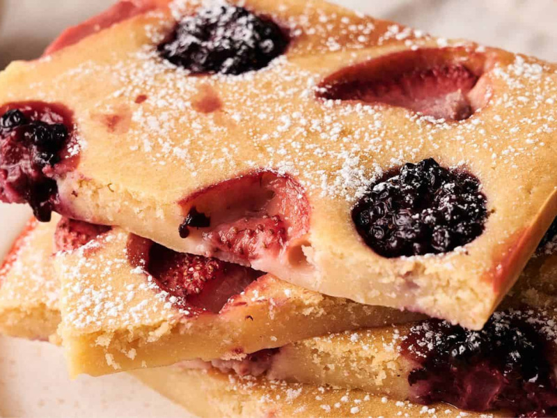 Three rectangular slices of baked fruit-studded cake, topped with powdered sugar, are stacked on a white plate.