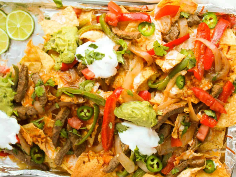 A tray of loaded nachos topped with sliced beef, red and green peppers, jalapeños, guacamole, sour cream, and lime wedges on the side.