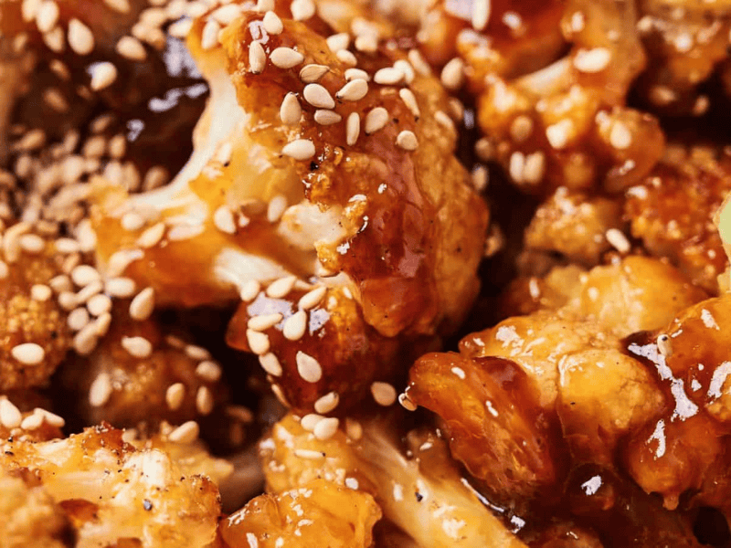 Close-up of Sticky Sesame Cauliflower: crispy florets coated in a glossy sauce, garnished with sesame seeds and sliced green onions.
