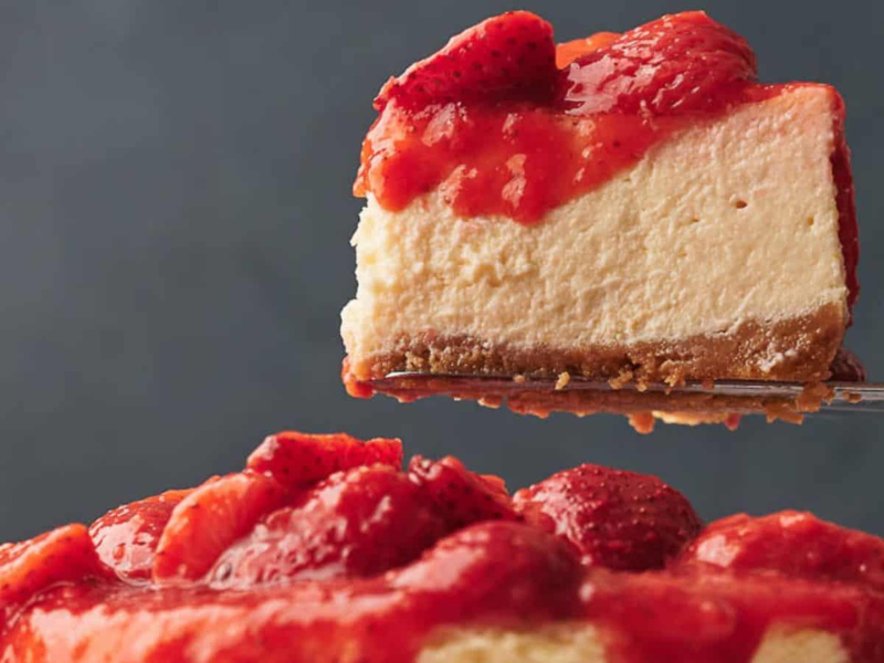 A slice of strawberry-topped cheesecake is being lifted above the rest of the cake, showing a graham cracker crust and creamy filling.
