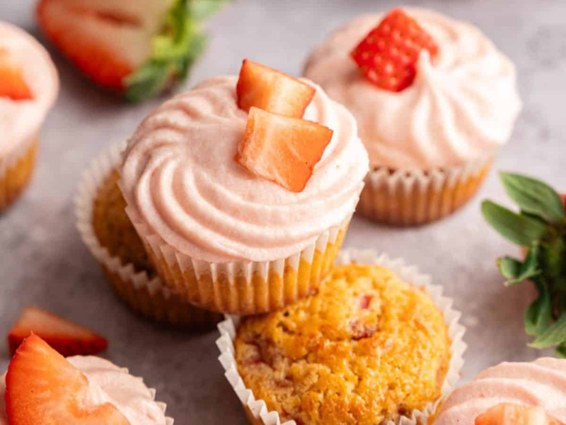Several cupcakes topped with pink strawberry frosting and fresh strawberry pieces are arranged on a light surface, with whole and sliced strawberries nearby.