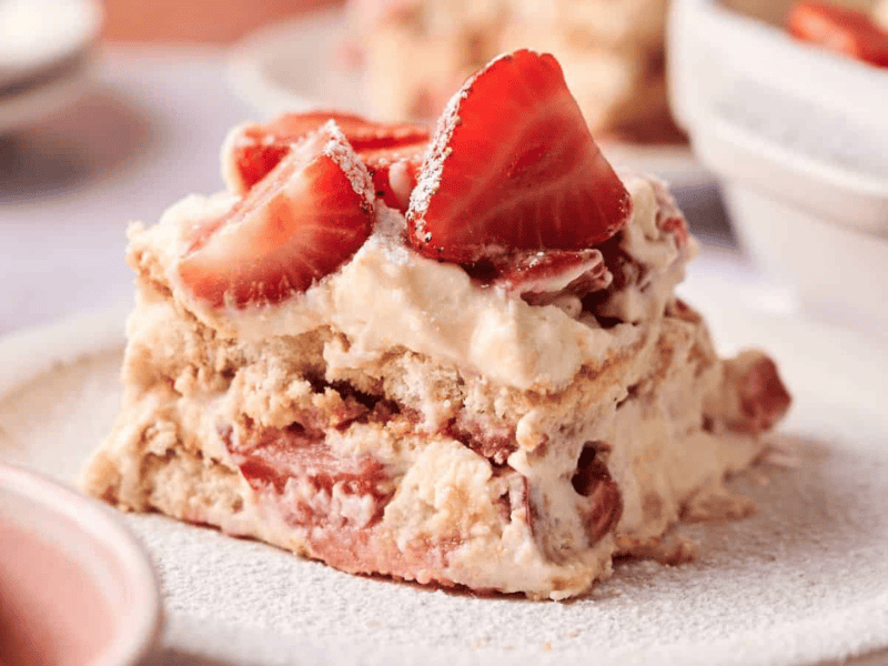 A slice of strawberry tiramisu with layers of cream, ladyfingers, and fresh strawberries is served on a white plate.