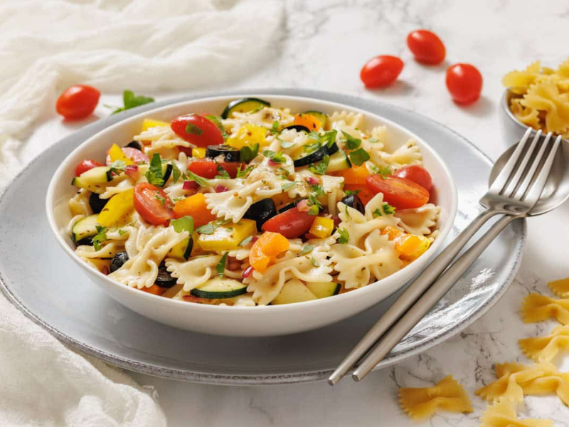 Bowl of bowtie pasta salad with cherry tomatoes, zucchini, bell peppers, and herbs, served on a plate with a fork and spoon.