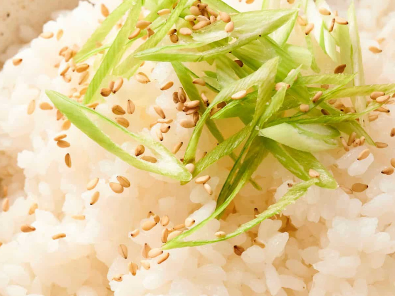 A bowl of steamed white rice topped with sliced green onions and sprinkled with sesame seeds.