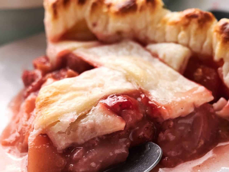 Close-up of a slice of fruit pie with a golden, flaky crust. The filling is a mix of pink and red fruit, slightly oozing out onto the plate.