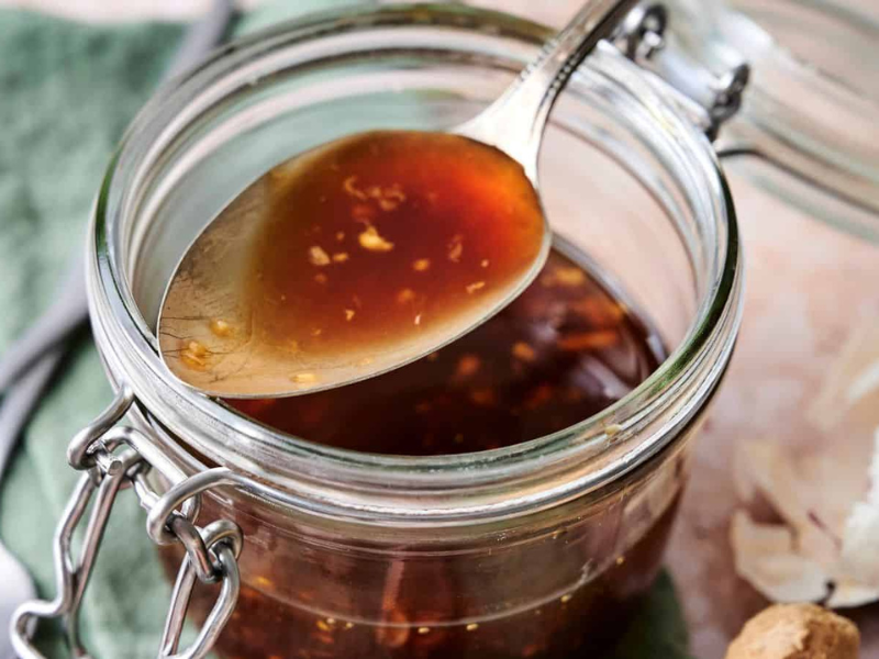 A spoon of brown liquid sauce with garlic bits is held above an open glass jar. A green cloth and garlic cloves are in the background.