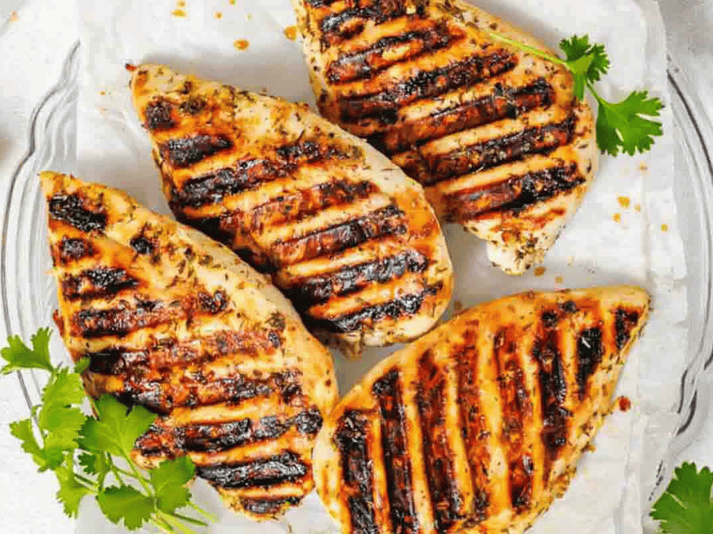 Four grilled chicken breasts with dark char marks are arranged on a white plate, garnished with fresh cilantro.