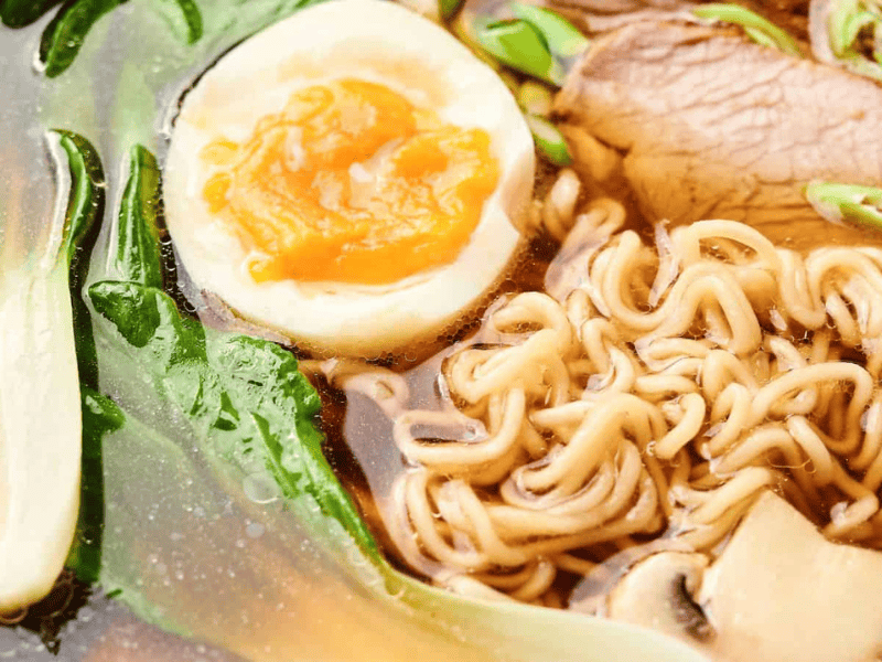 Close-up of a bowl of ramen with noodles, a halved boiled egg, sliced meat, green leafy vegetables, and mushrooms in broth.
