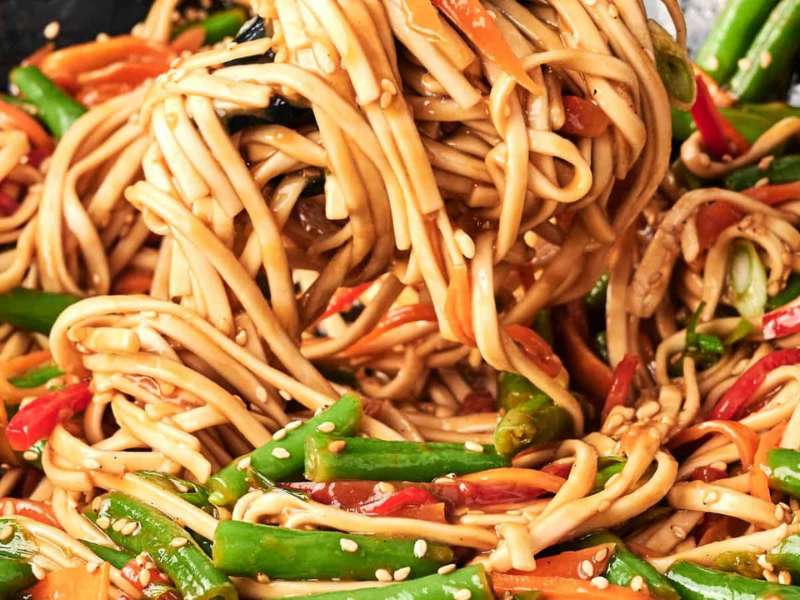A close-up of stir-fried noodles with green beans, red bell peppers, and carrots, garnished with sesame seeds.