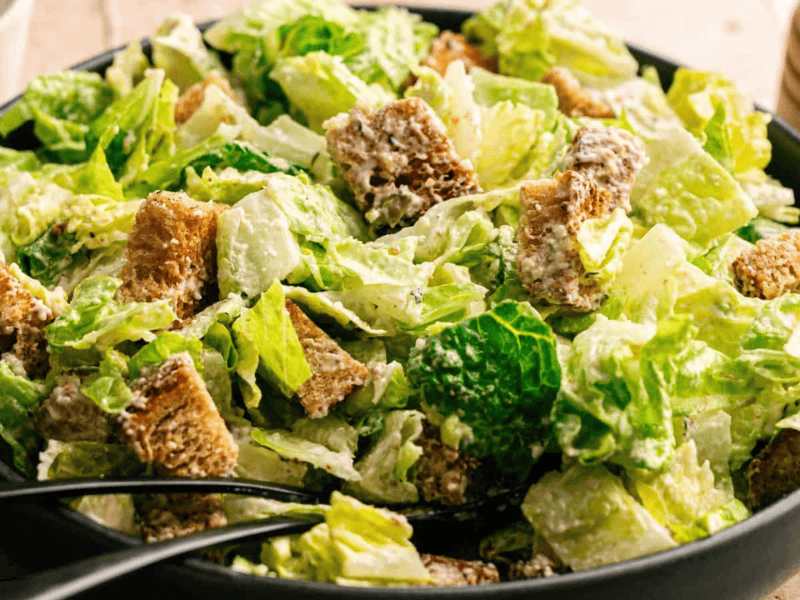A bowl of Caesar salad with romaine lettuce, croutons, and creamy dressing, with serving tongs resting in the salad.