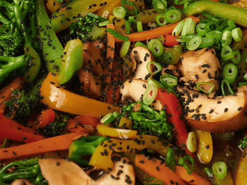 Close-up of stir-fried vegetables including broccoli, bell peppers, carrots, mushrooms, and green onions, topped with black sesame seeds.