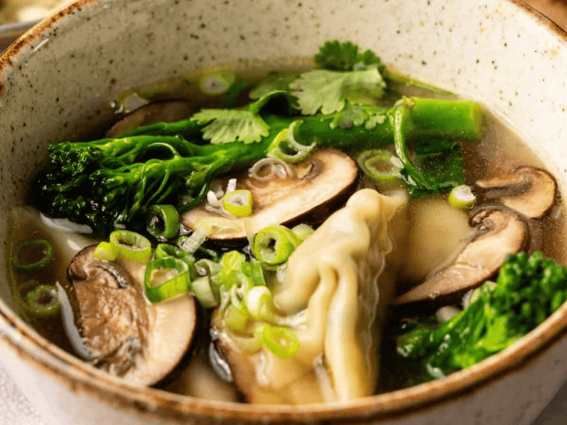 A ceramic bowl of clear soup containing dumplings, sliced mushrooms, broccolini, chopped green onions, and cilantro.