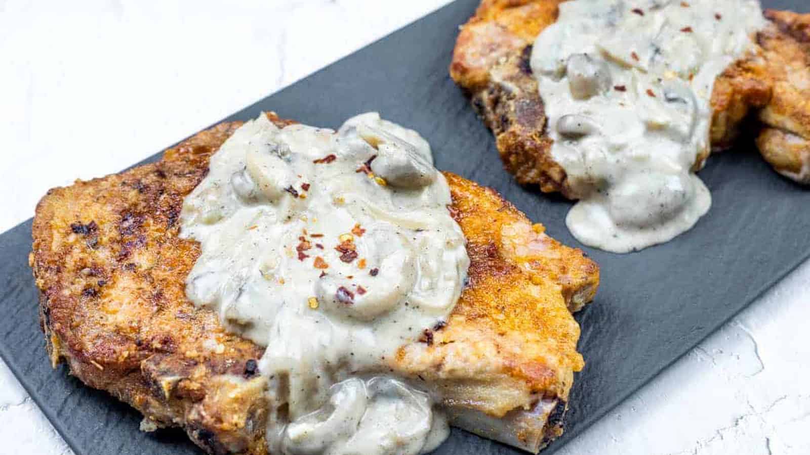 Two Smothered Pork Chops with Onions & Mushrooms on a black rectangular plate.