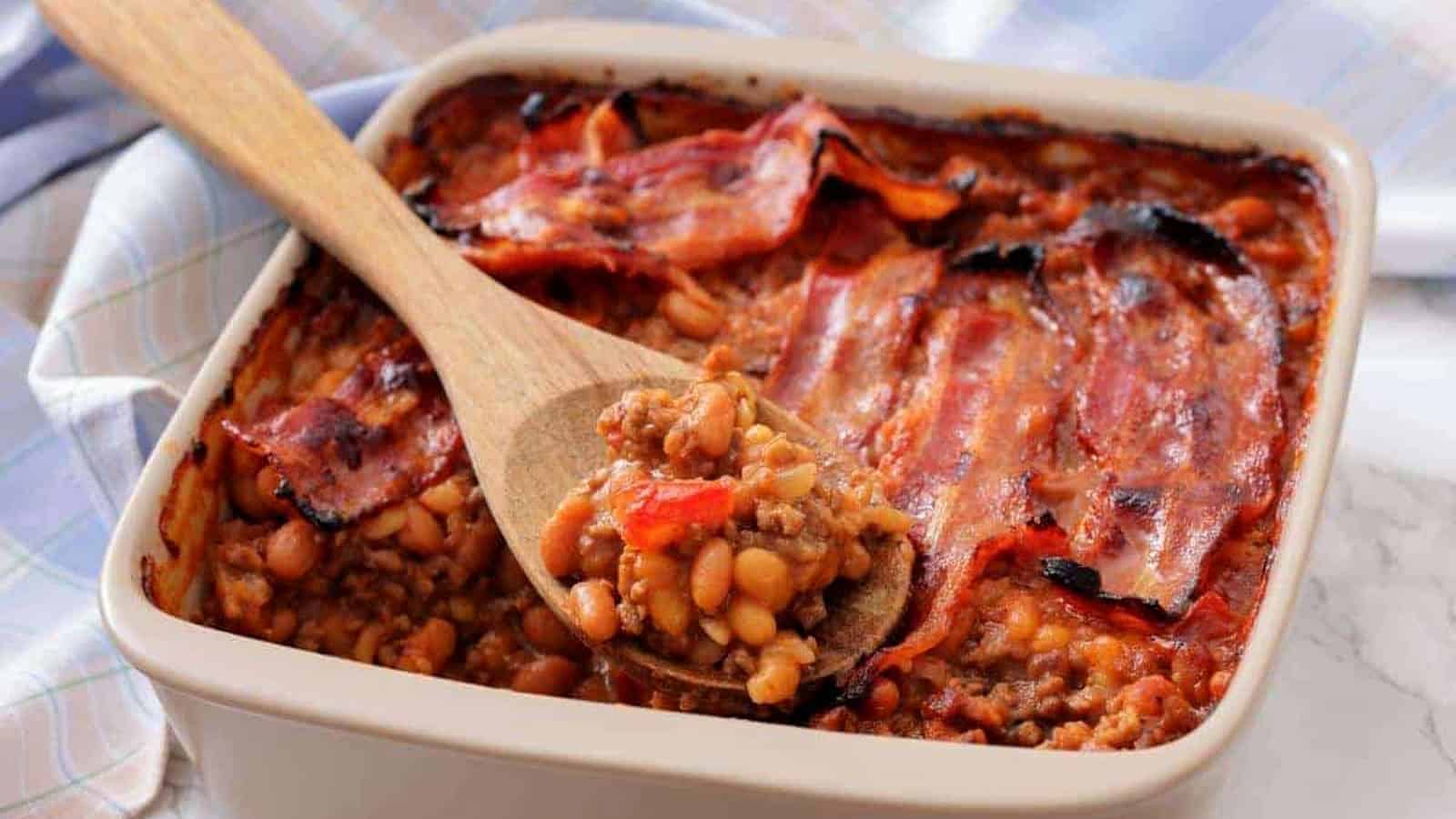A square baking dish filled with Southern Ground Beef & Baked Bean Casserole and topped with strips of cooked bacon; a wooden spoon holds a serving.